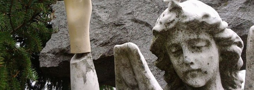 A weathered stone angel stands before a granite cross; one hand has been replaced by a smooth, plastic mannequin hand raised in greeting.
