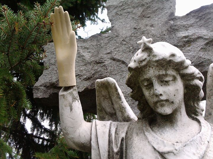 A weathered stone angel stands before a granite cross; one hand has been replaced by a smooth, plastic mannequin hand raised in greeting.